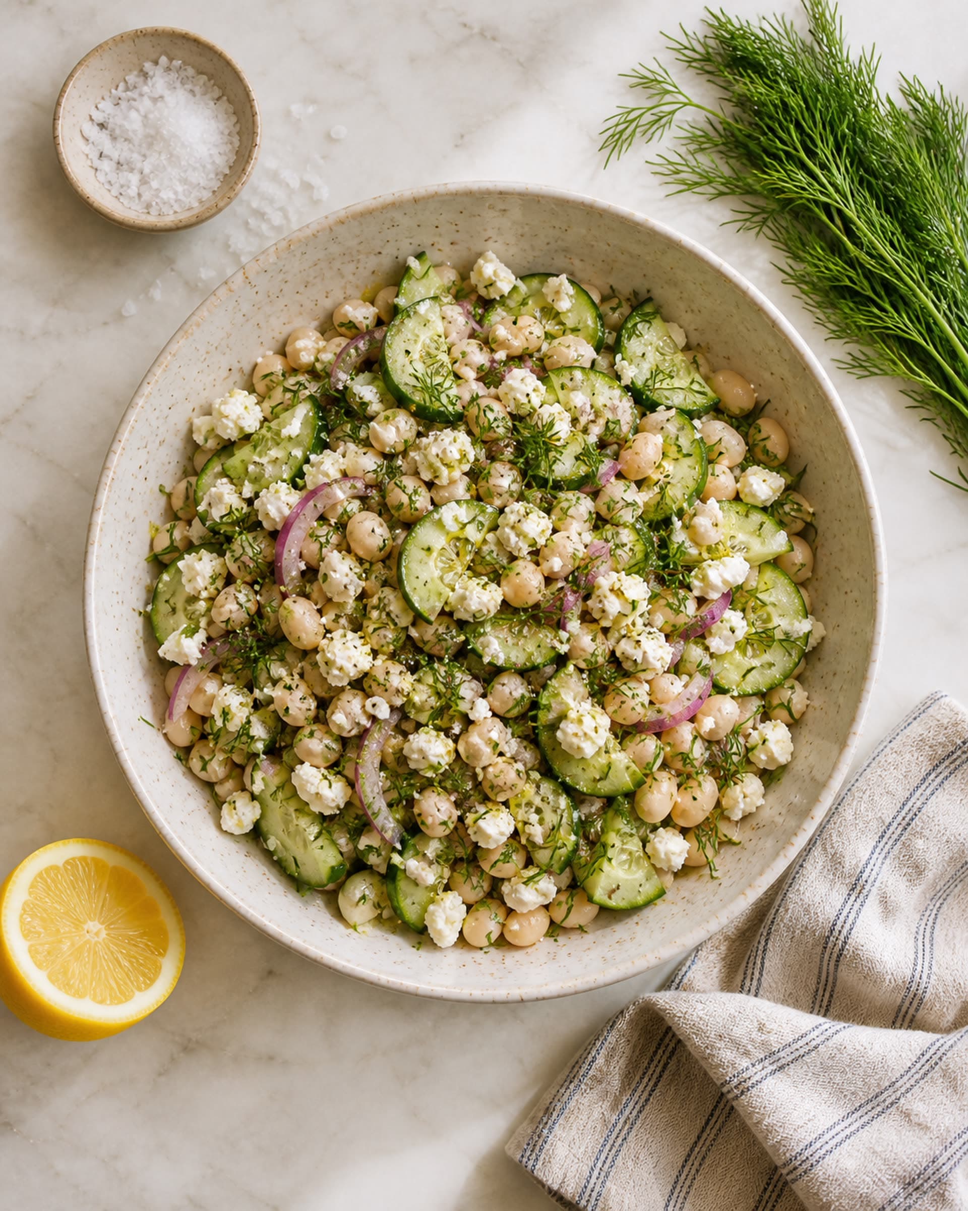 A near-overhead view of a wide shallow cream-speckled stoneware bowl on a white marble counter, filled with a cold salad of creamy white cannellini beans, thin half-moons of crisp cucumber, slivers of red onion, a generous scatter of crumbled white feta, a lot of finely chopped fresh dill, and a few cracks of black pepper, all glossy with a lemon and olive oil dressing. A halved lemon, a small bunch of fresh dill, a small dish of flaky salt, and a folded striped cream-and-blue linen tea towel rest on the marble beside the bowl. Soft bright midday light.