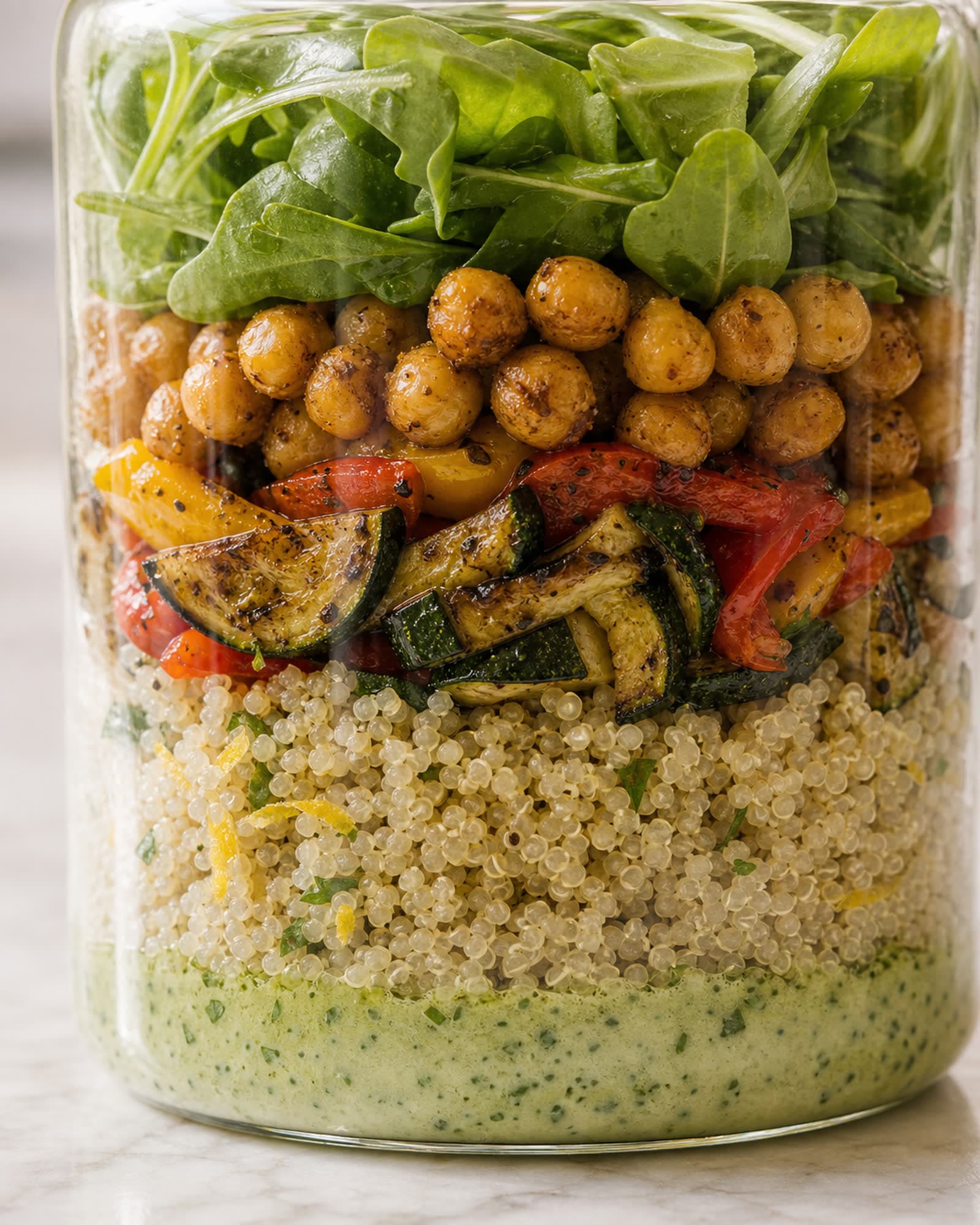 A close macro looking into the side of a glass meal-prep jar, showing the distinct layers: green herb dressing at the bottom, fluffy quinoa, roasted zucchini and peppers, crispy chickpeas, and fresh greens on top.