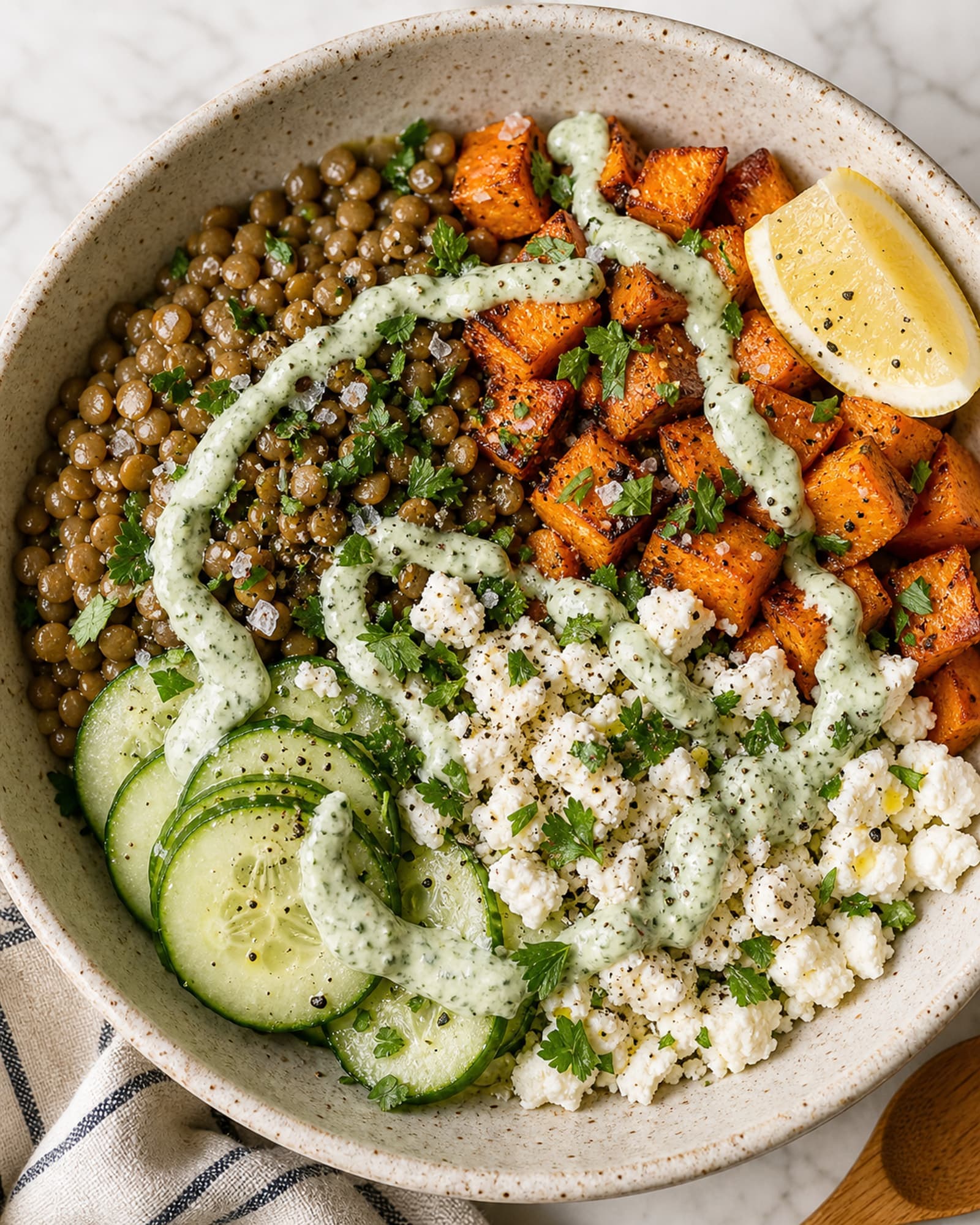 Near-top-down macro of one assembled lentil and feta meal-prep bowl in a wide cream-speckled stoneware bowl on a white marble counter: a generous pile of warm French green lentils glossy with olive oil at one side, a section of cubed roasted sweet potatoes with caramelized edges at another, a small fan of thinly sliced cucumber rounds, a generous crumble of white feta, scattered chopped fresh parsley across everything, a small wedge of lemon perched on the rim, a generous swirl of pale-green herb-yogurt-tahini dressing drizzled across the top, a flake of flaky salt, and visible cracks of black pepper.