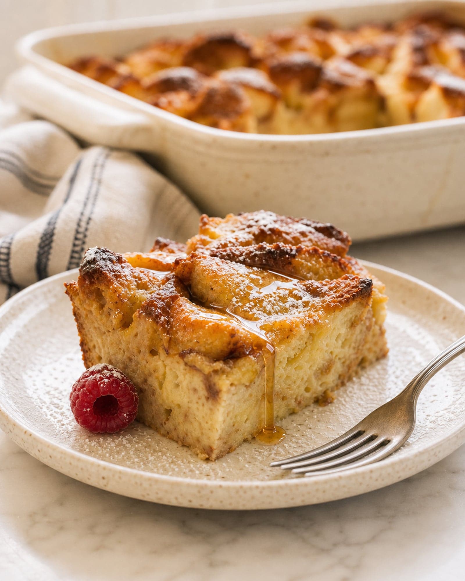 A close-up of one corner square of french toast casserole on a speckled plate, crisp golden peaks on top and a soft custardy interior at the cut face, with a thread of maple syrup dripping down and a raspberry beside it.
