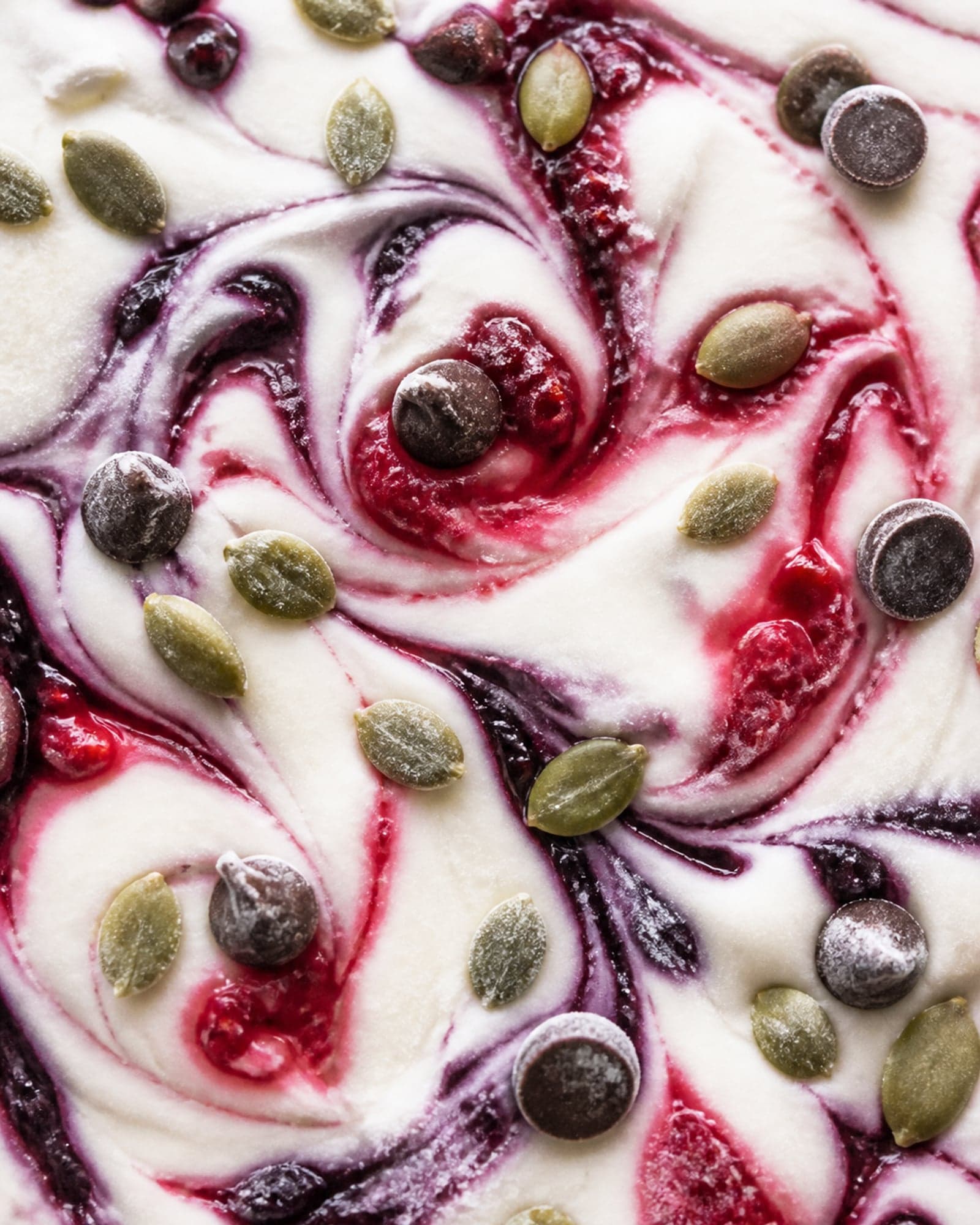 A close macro of the protein bark: creamy skyr dragged into swirls with red raspberry and blueberry, scattered with pumpkin seeds and dark chocolate, with a frosty frozen look.