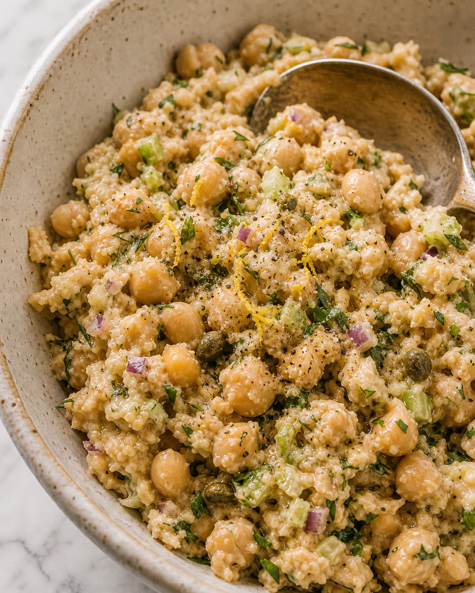 A close macro of the smashed chickpea salad, showing half-mashed and half-whole chickpeas, flecks of fresh dill and parsley, small dice of celery and red onion, and a creamy lemony bind, with a spoon resting in it.