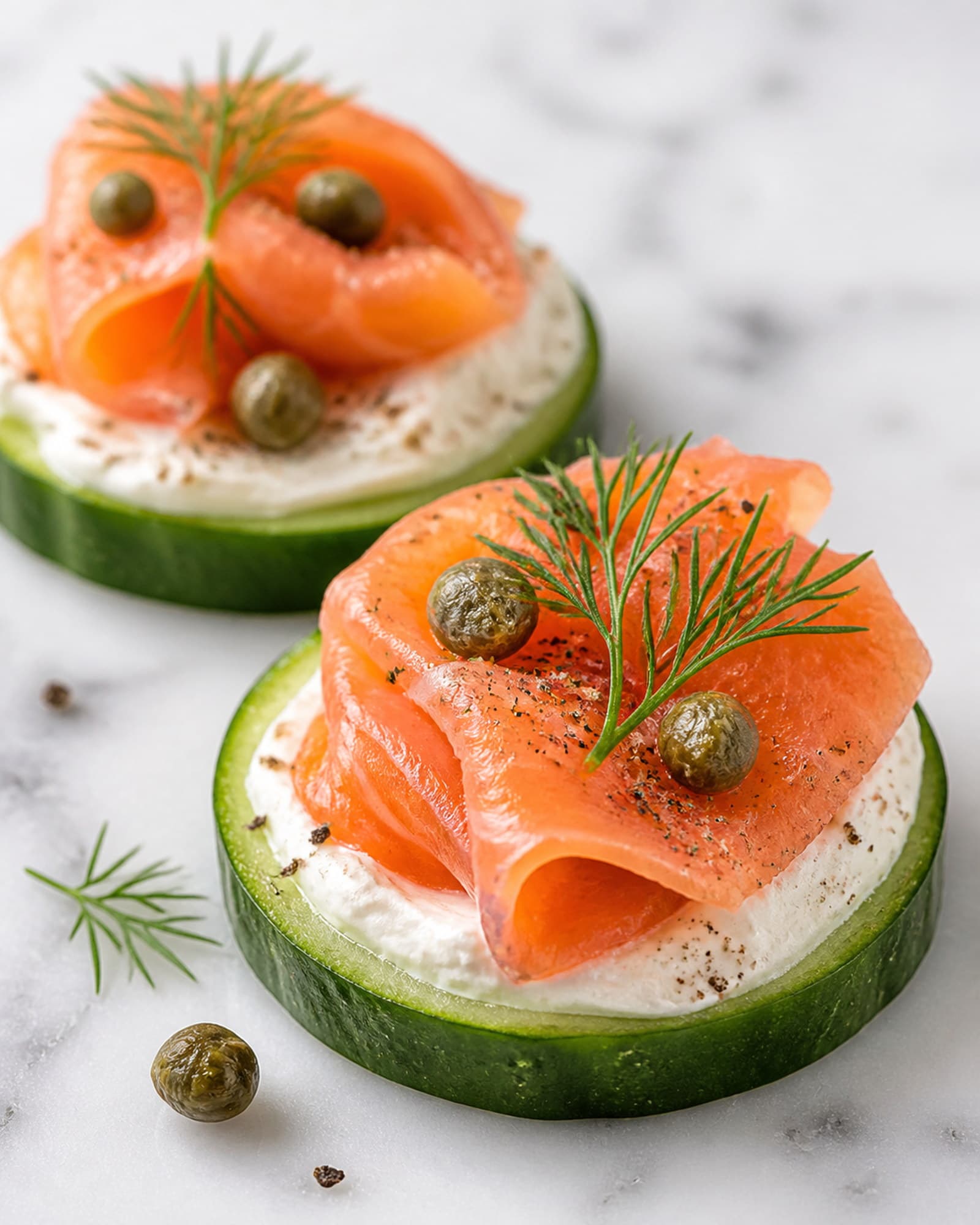 A close macro of a finished smoked salmon cucumber bite: a ruffled curl of salmon over lemony cream cheese on a cucumber round, topped with green capers, a frond of dill, and cracked pepper.