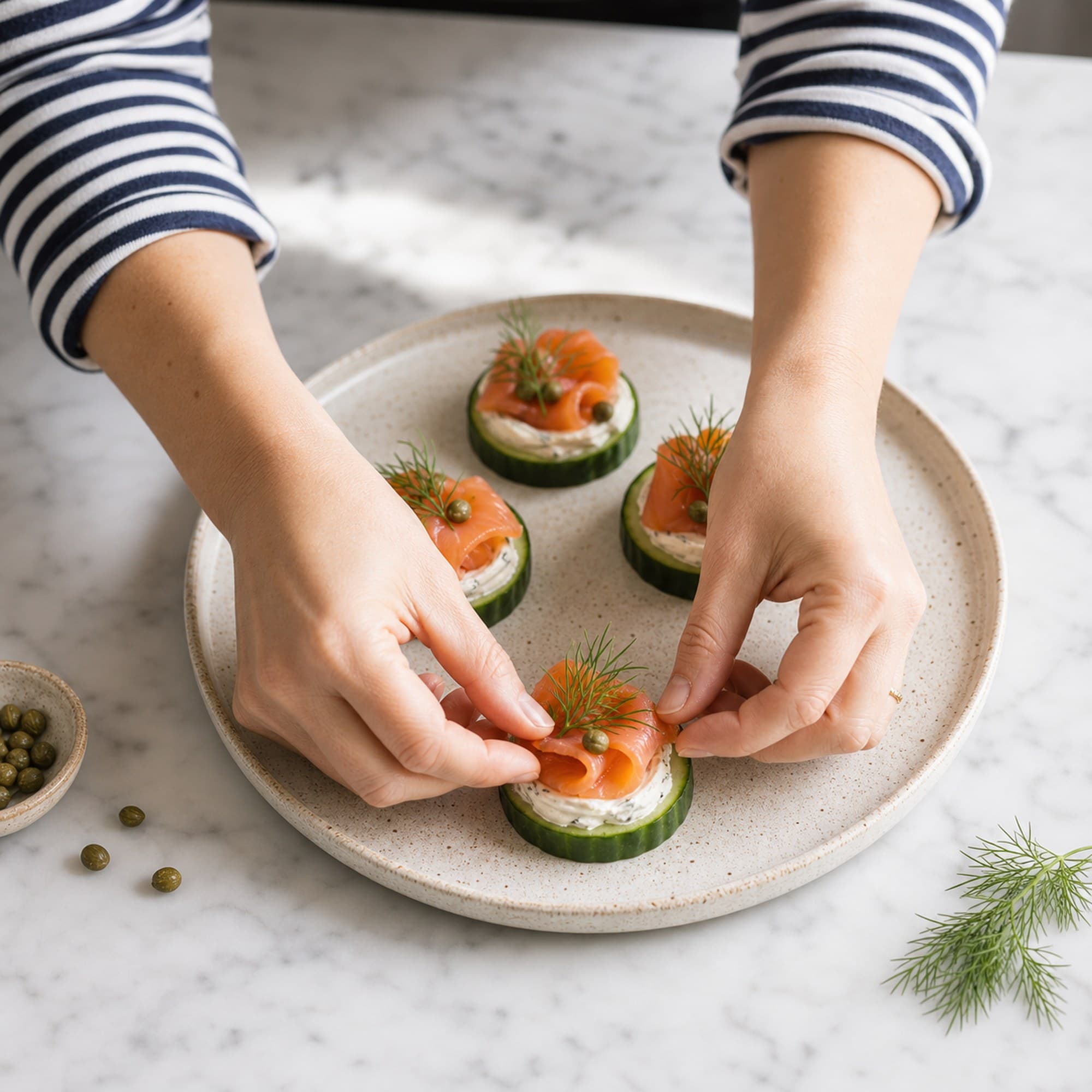 A pair of hands in a navy striped sleeve placing a frond of fresh dill onto a finished smoked salmon cucumber bite on a plate, on a marble counter.