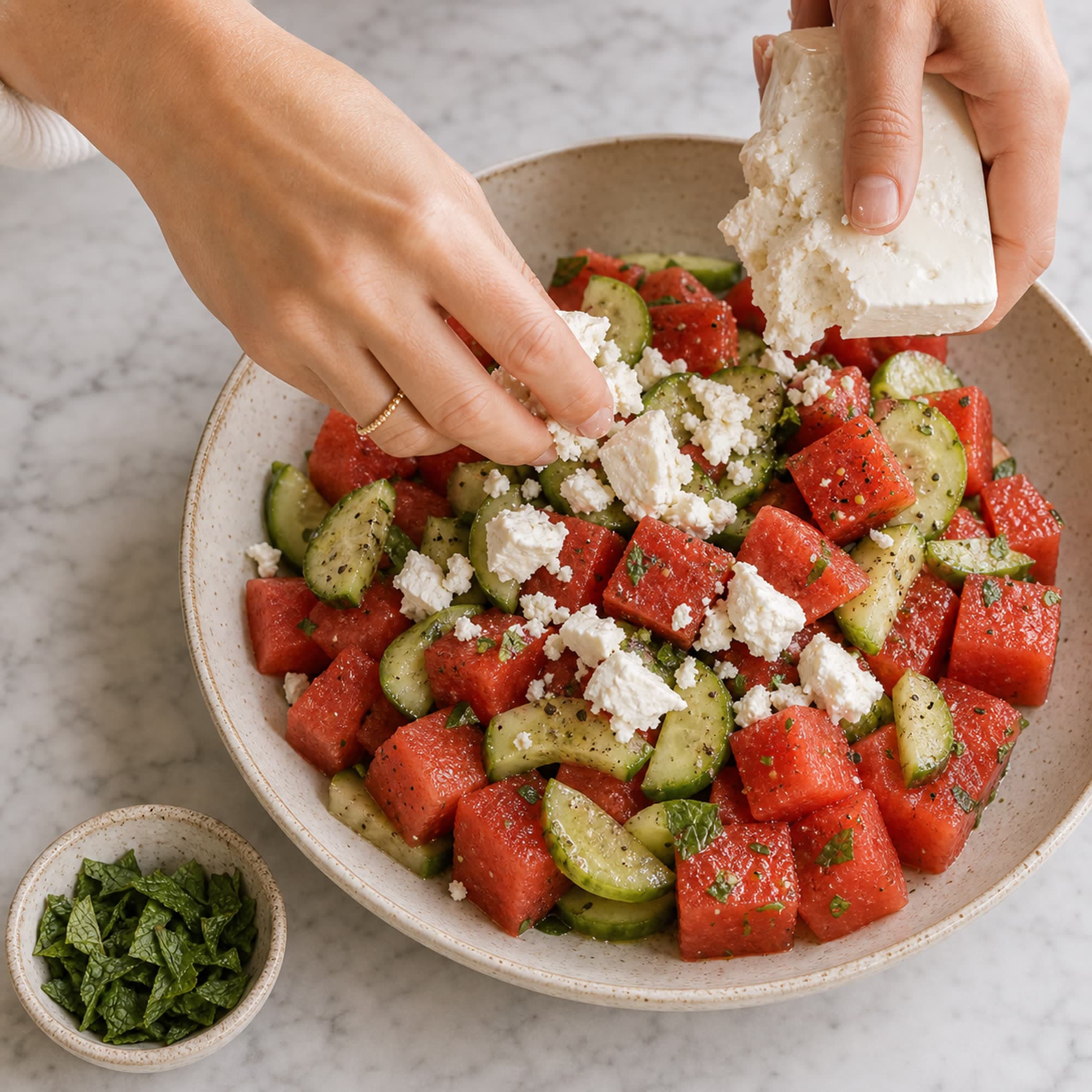 Hands breaking a block of feta into rough crumbles over a wide bowl of watermelon cubes and cucumber half-moons on a marble counter, with a small pinch bowl of torn mint waiting beside it.
