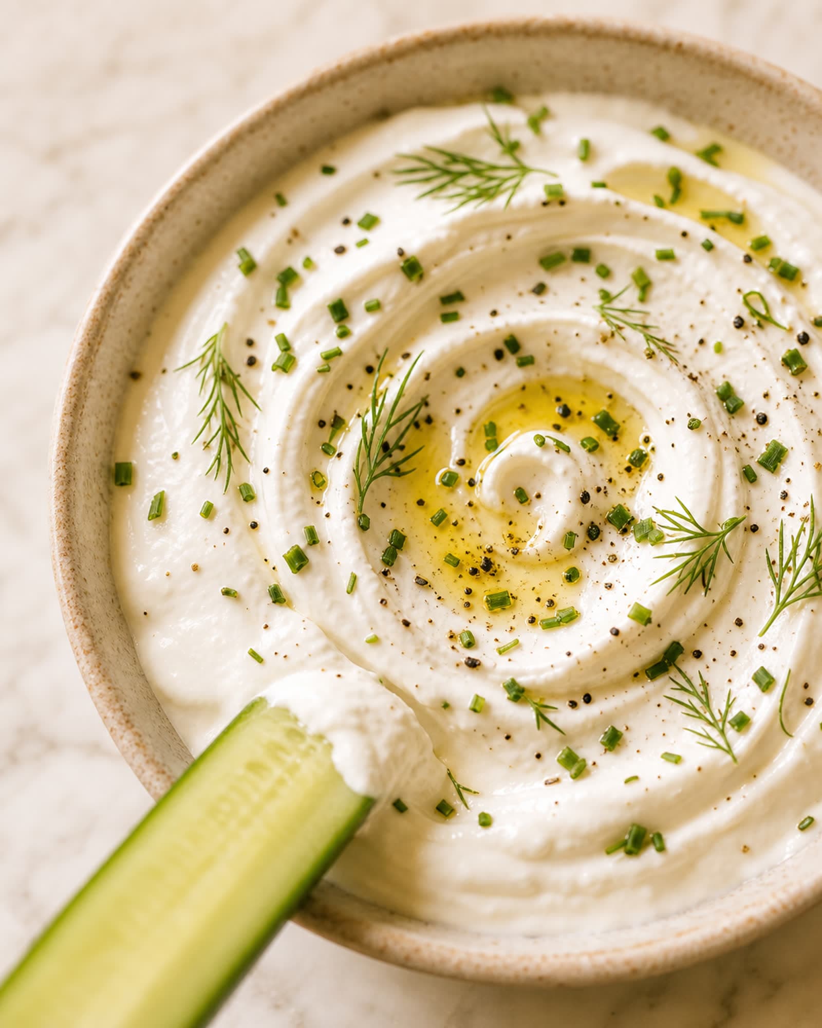 A close macro of the silky whipped cottage cheese dip, olive oil pooling in the swirl, scattered with chopped chives and dill and cracked pepper, with one cucumber spear dipping in.