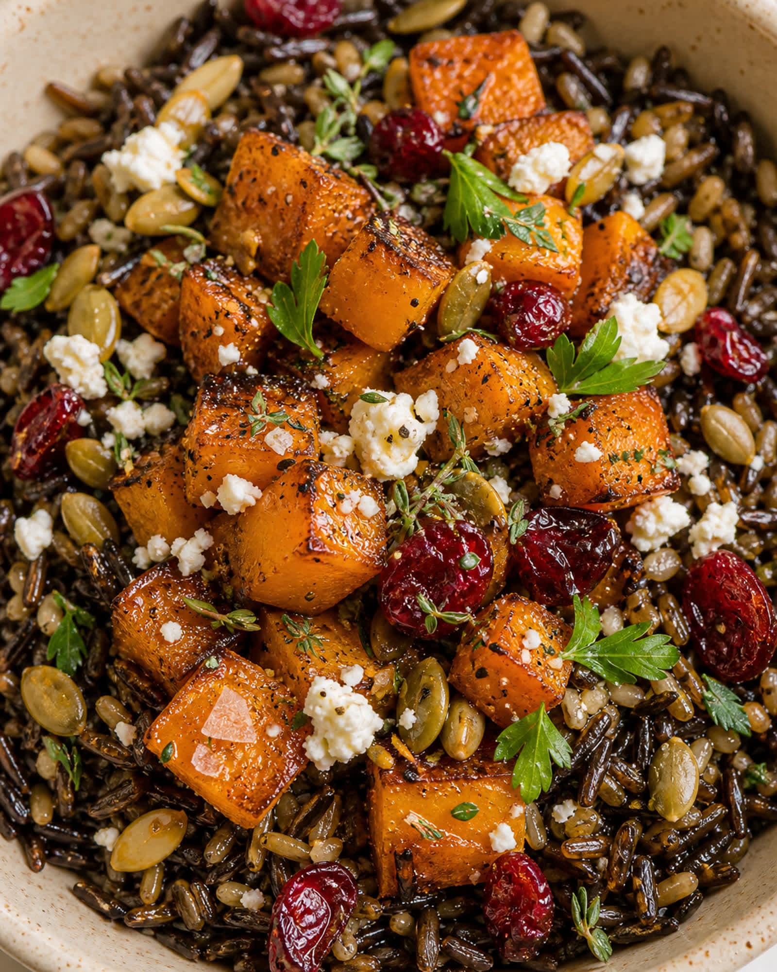 Macro top-down close-up of an assembled wild rice and squash bowl surface, showing dark nutty wild rice with split grains, vibrant orange caramelized butternut squash cubes with charred edges, bright red dried cranberries scattered through, white crumbled feta cheese, golden green-rimmed pumpkin seeds, small parsley leaves and tiny thyme sprigs, with cracked black pepper across the squash and a flake of flaky salt visible. The cream-speckled stoneware bowl edge sits softly out of focus at the corners.