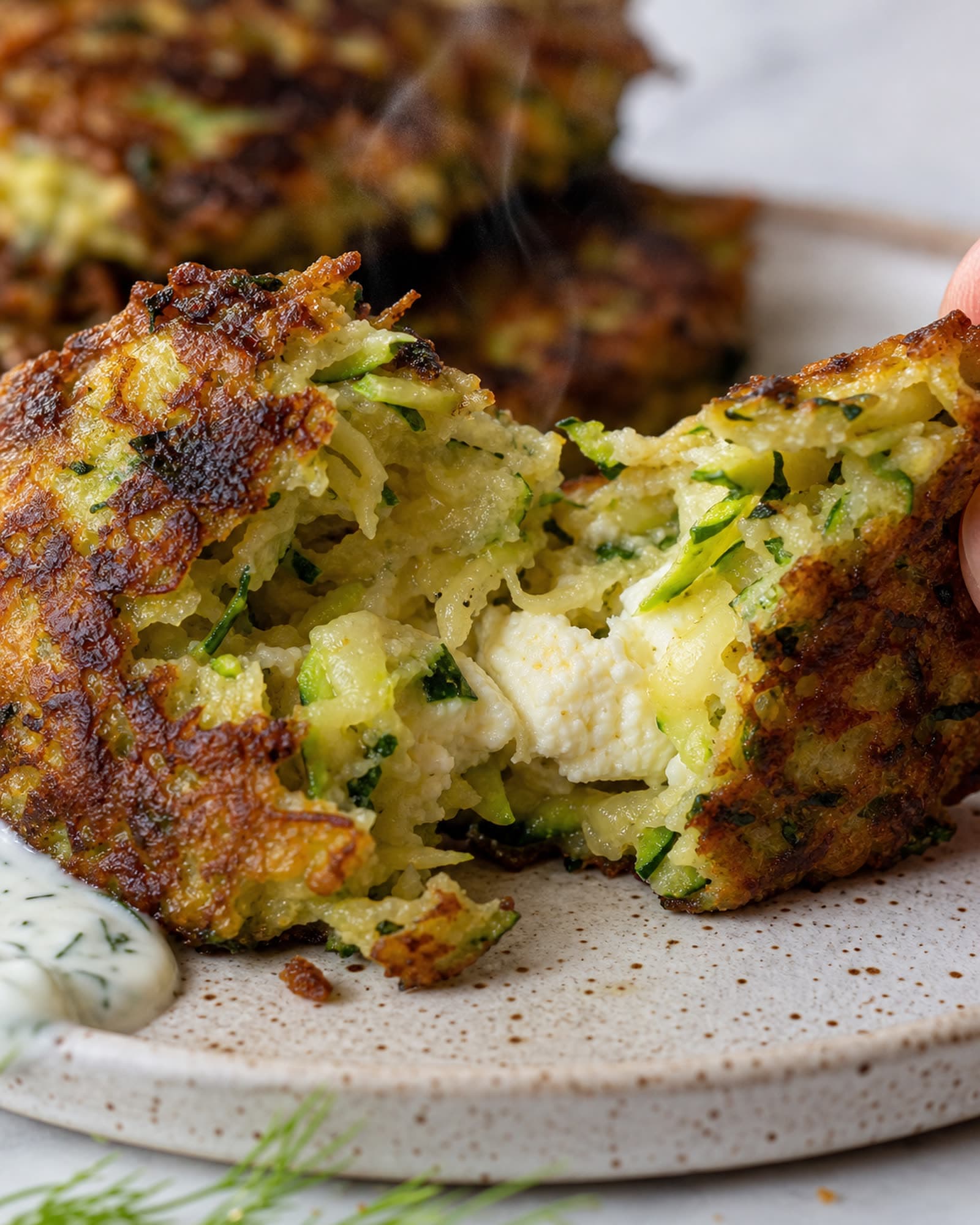 A zucchini fritter torn in half showing the tender green-flecked interior with a pocket of melted feta, steam rising from the crisp golden crust, more fritters blurred behind on a speckled plate.